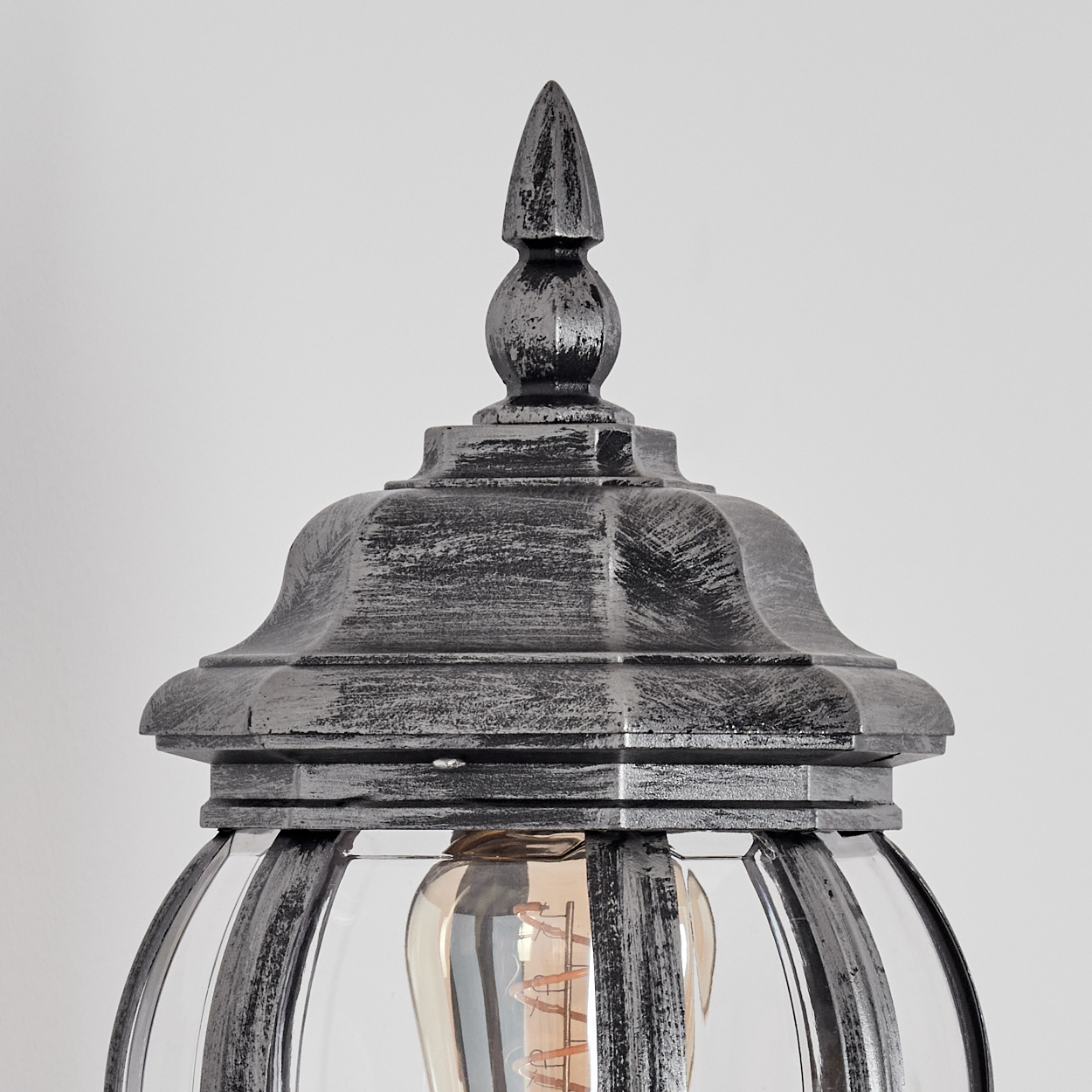 Nahaufnahme einer dekorativen Lampe. Der obere Teil ist metallisch grau mit einer spitzen Spitze. Sie hat ein Glasgehäuse mit einer sichtbaren Glühbirne.