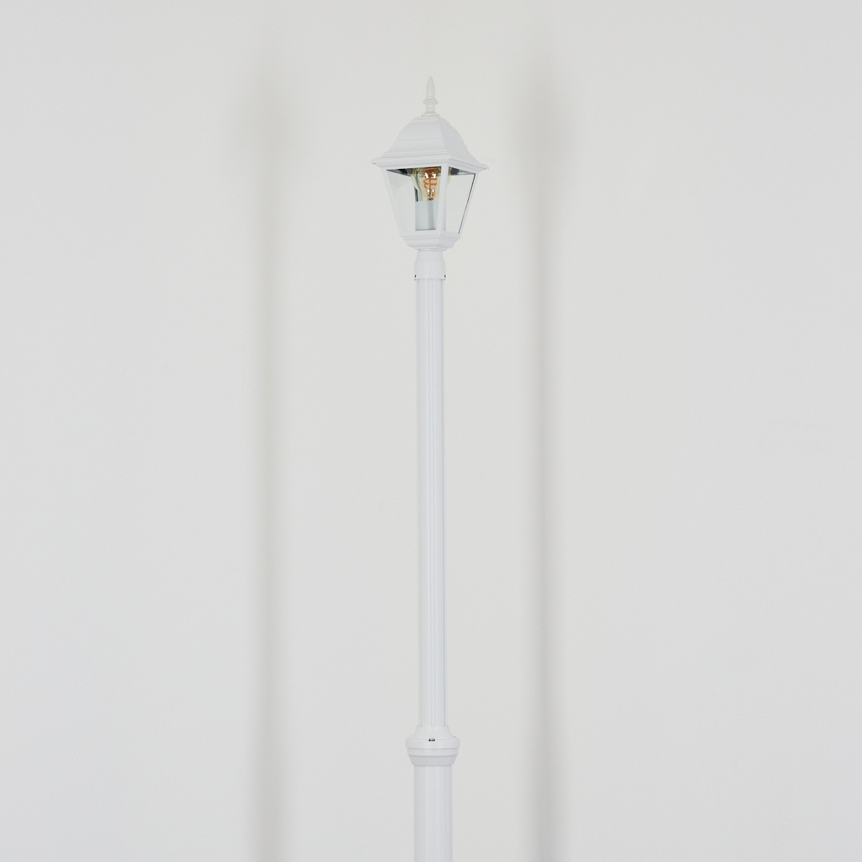 Eine weiße Straßenlaterne steht vor einer weißen Wand. Die Lampe hat ein gläsernes Gehäuse und eine dekorative Spitze.