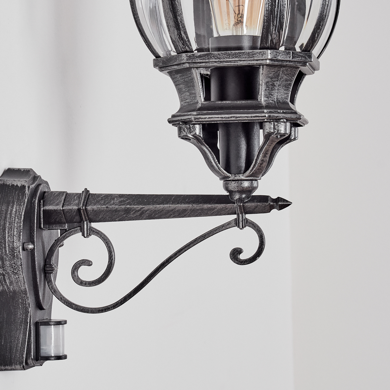 Eine dekorative Außenlampe an einer weißen Wand. Die Lampe hat eine silberne und schwarze Oberfläche mit einem Glasgehäuse und einem Bewegungssensor.
