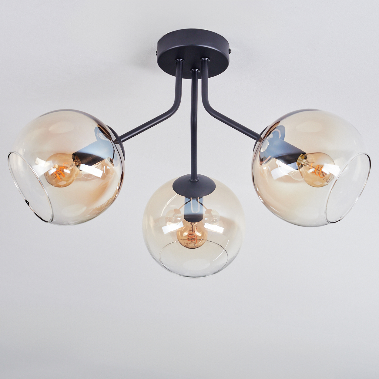 Eine moderne Deckenleuchte mit drei kugelförmigen, bernsteinfarbenen Glasschirmen, jede mit einer Glühbirne, an schwarzen Metallarmen aufgehängt.