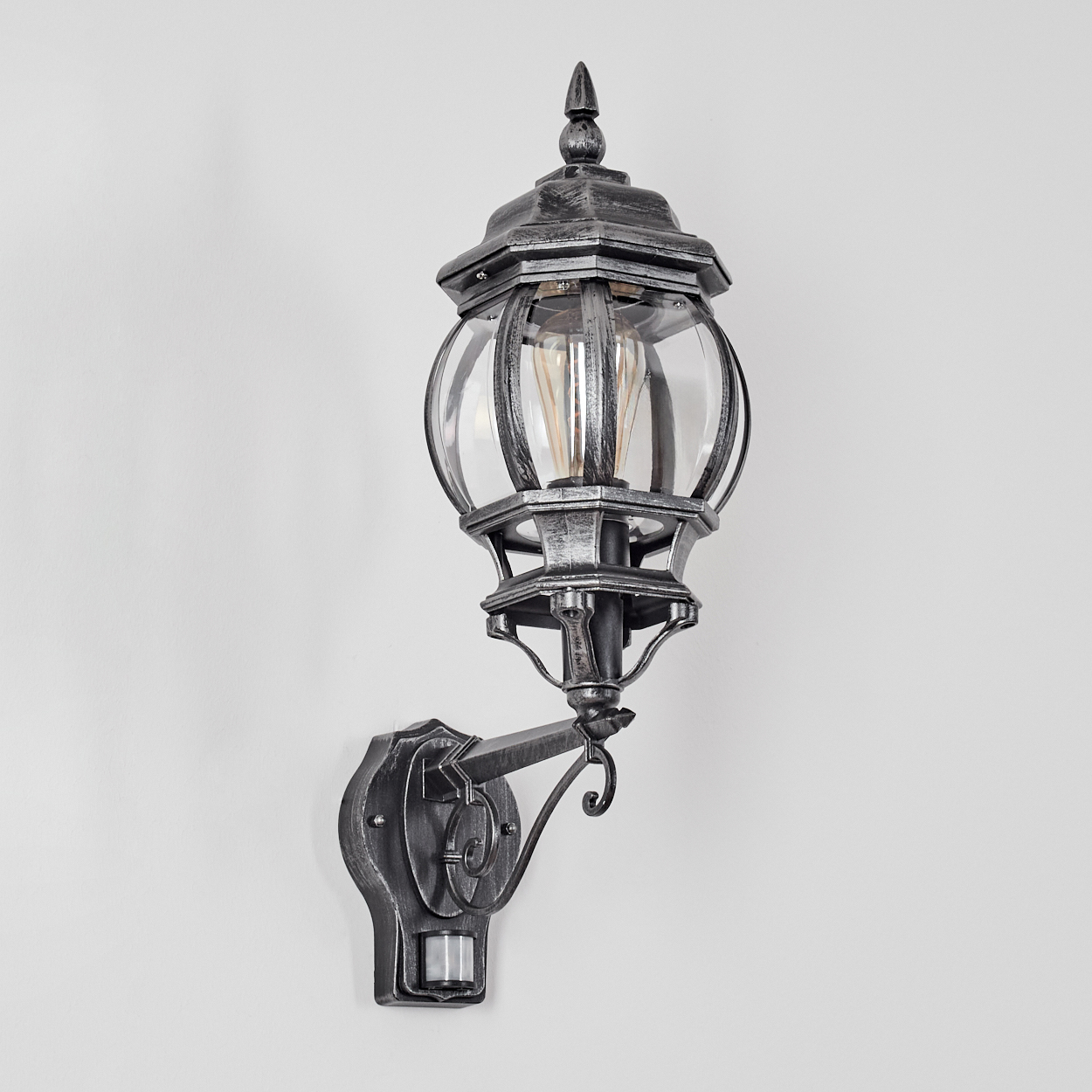 Eine dekorative Außenlampe, silbergrau, an einer weißen Wand montiert. Sie hat ein Glasgehäuse und einen Bewegungssensor.