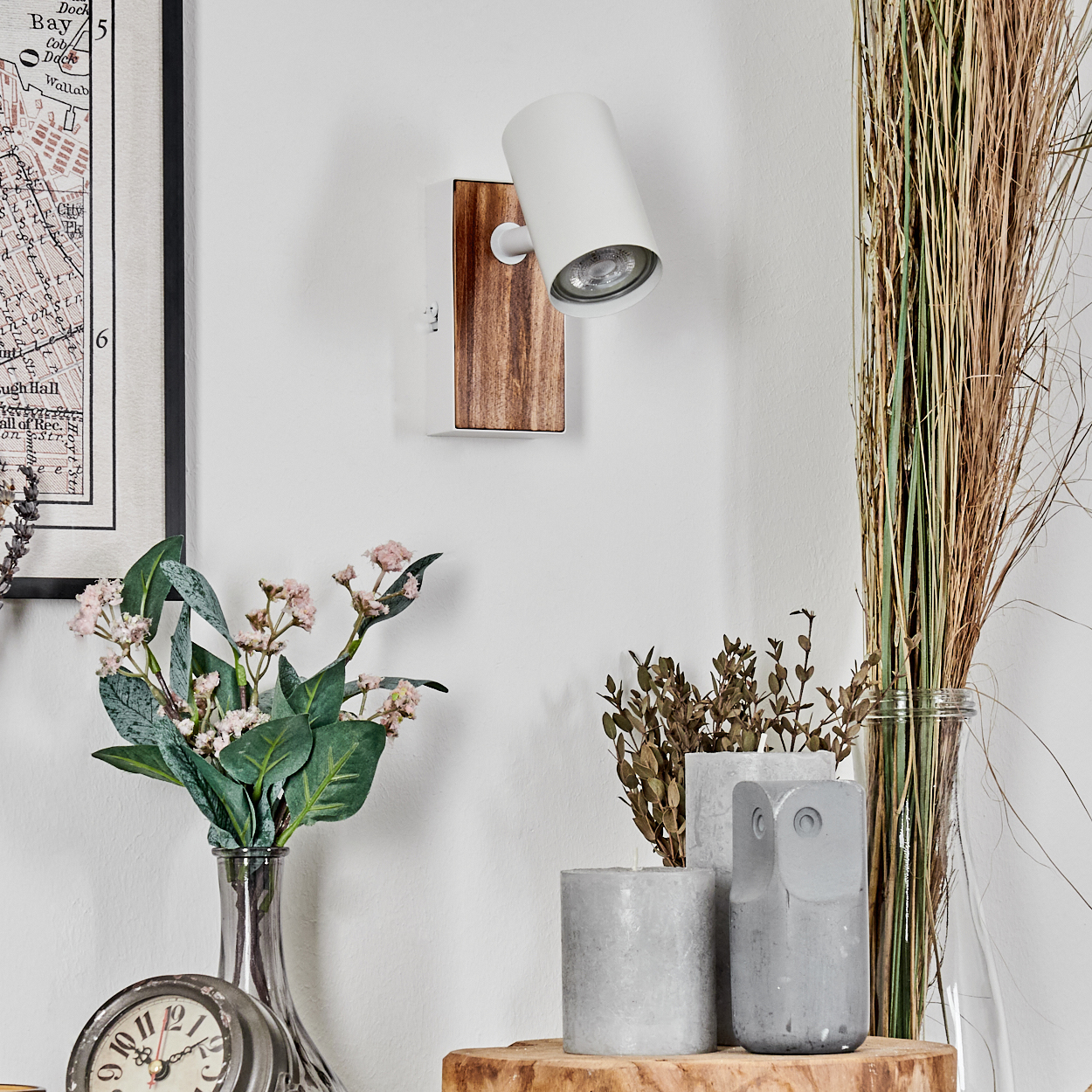 Eine weiße Lampe ist auf einer Holzbasis an einer weißen Wand montiert. Dekorative Elemente sind Blumen, eine Uhr und Ziergräser.