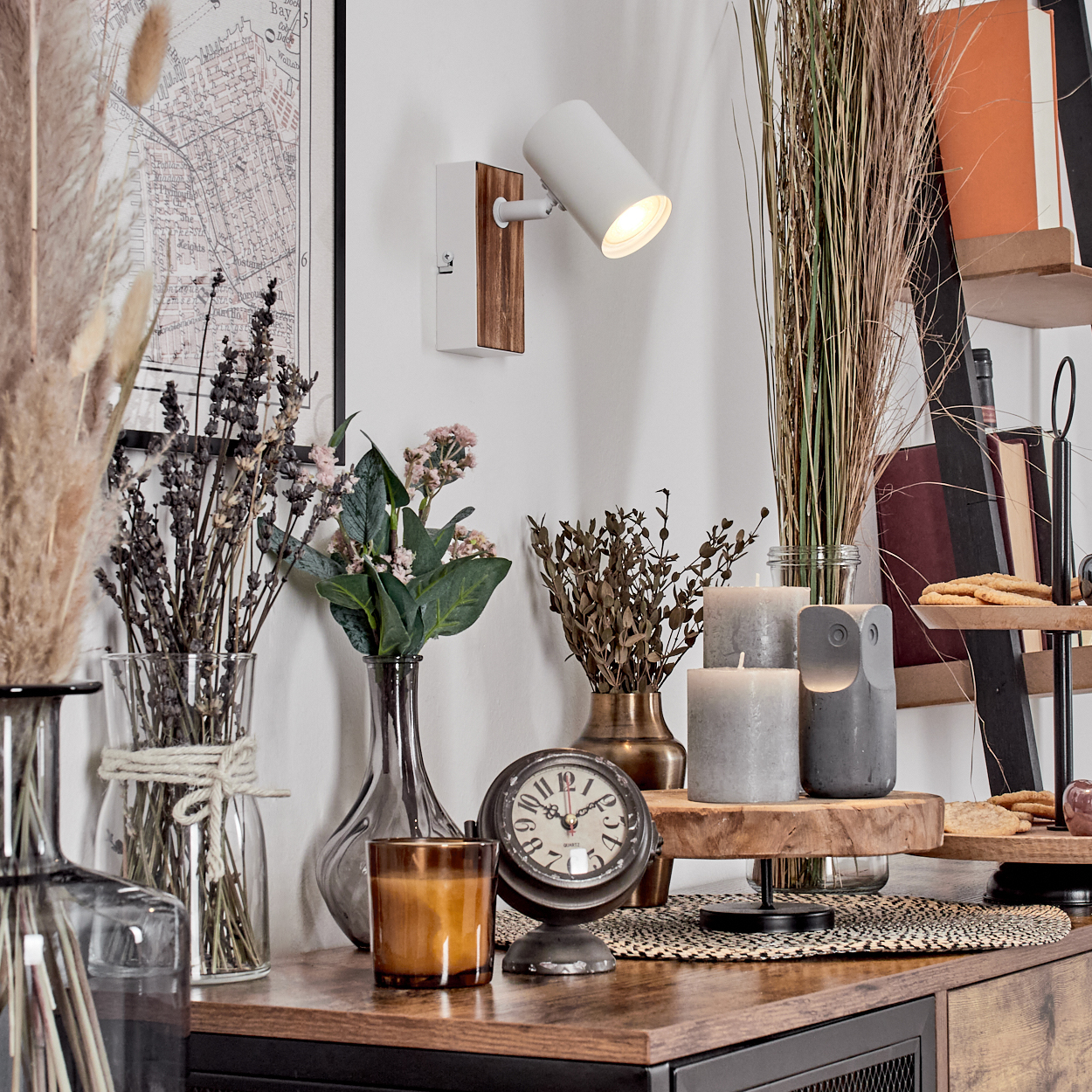 Eine dekorative Szene mit Trockenblumen, Kerzen, einer Uhr und einer Wandlampe auf einem Holztisch. Im Hintergrund ist eine Karte zu sehen.