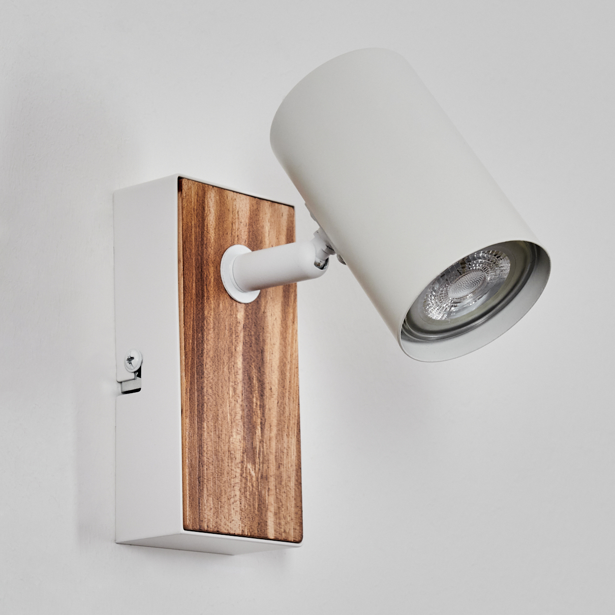 Eine weiße Wandleuchte mit Holzsockel. Die Lampe ist zylindrisch und angewinkelt. Die Basis ist rechteckig, und eine Schraube ist sichtbar.