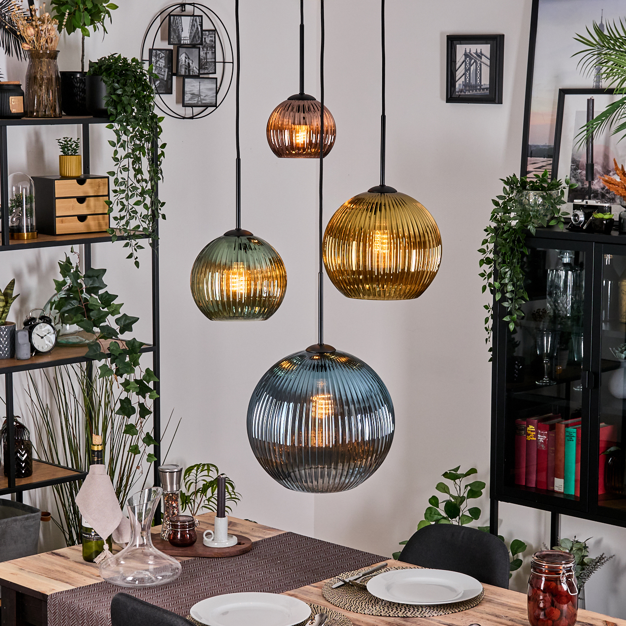 Vier bunte Pendelleuchten über einem Holztisch, der zum Abendessen gedeckt ist. Regale mit Pflanzen und Dekoration sind sichtbar.