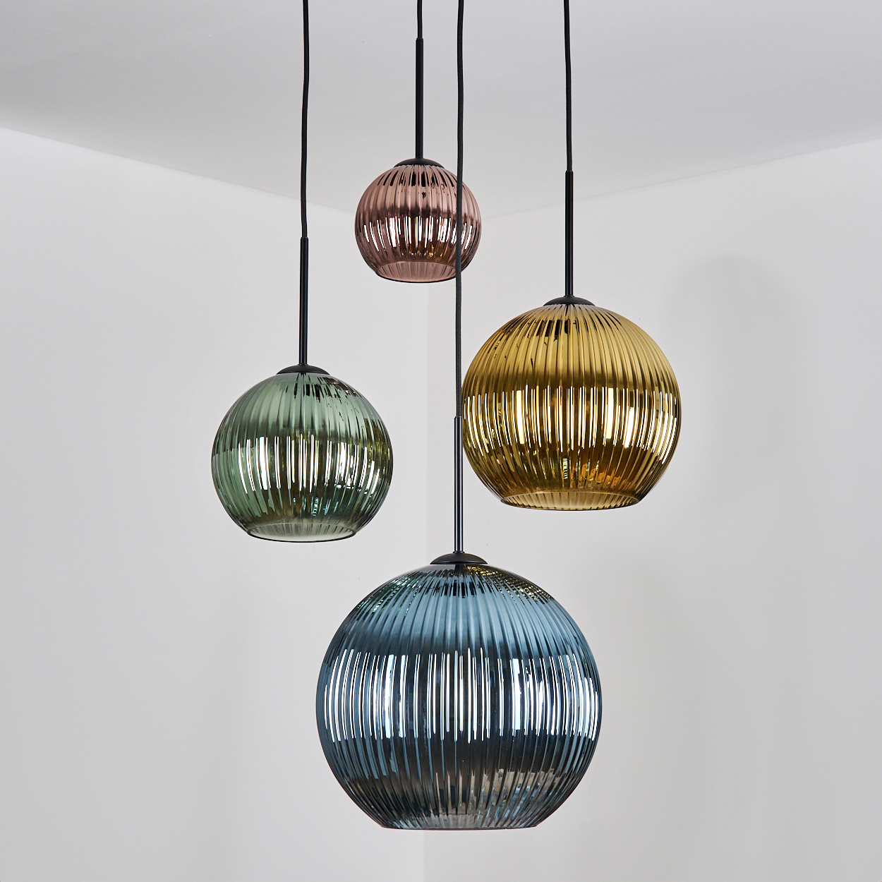 Vier kugelförmige, gerippte Pendelleuchten in Grün, Gold, Rosa und Blau hängen vor einer weißen Wand.