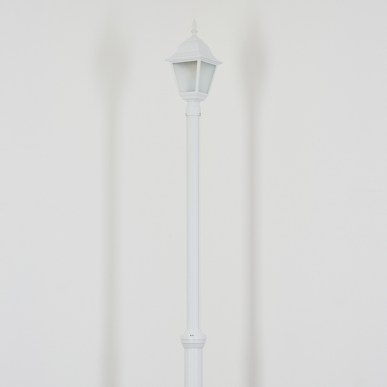 Eine weiße Straßenlaterne steht mittig vor einer weißen Wand. Die Lampe hat ein klassisches Design mit einem Laternenkopf.