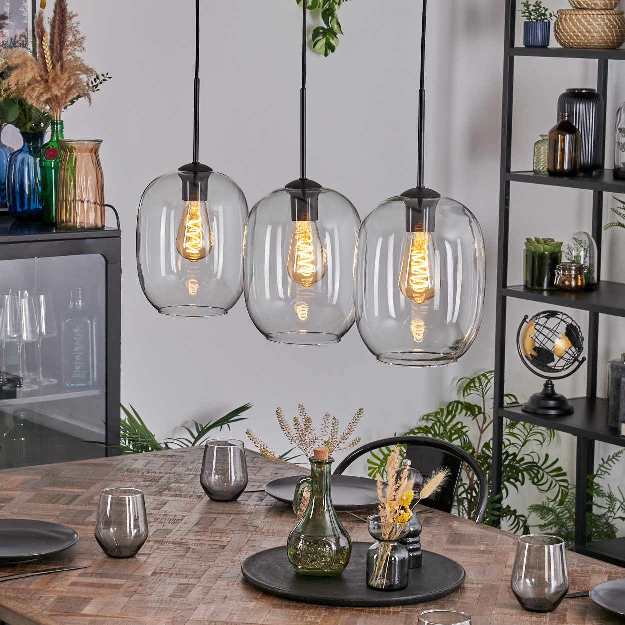 Drei Glas-Pendelleuchten über einem Holztisch, der für ein Essen gedeckt ist. Vasen und Gläser stehen auf dem Tisch, im Hintergrund Regale.