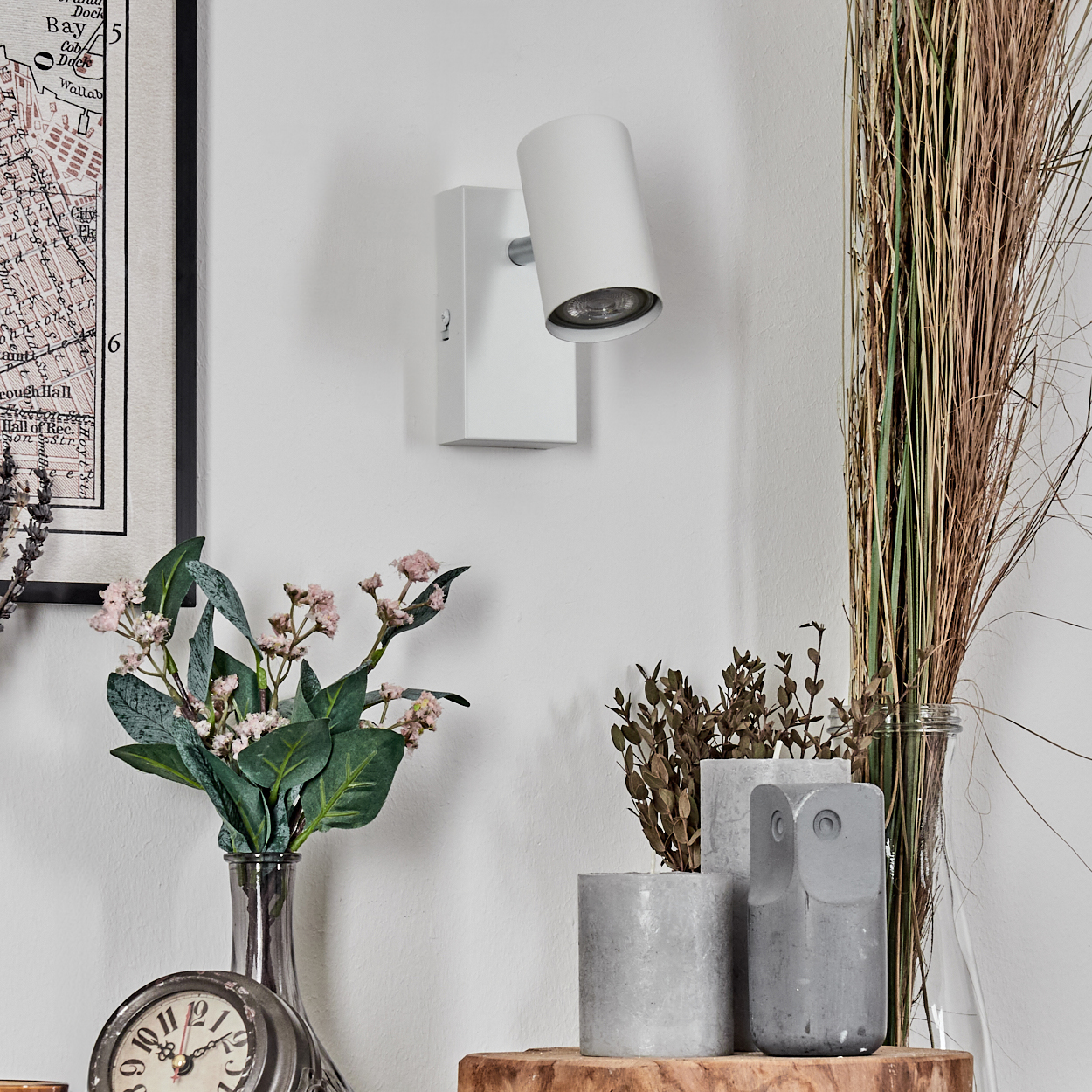 Eine weiße Wandleuchte ist an einer weißen Wand befestigt. Darunter befinden sich Dekorationen, darunter Pflanzen, eine Uhr und eine Vase.