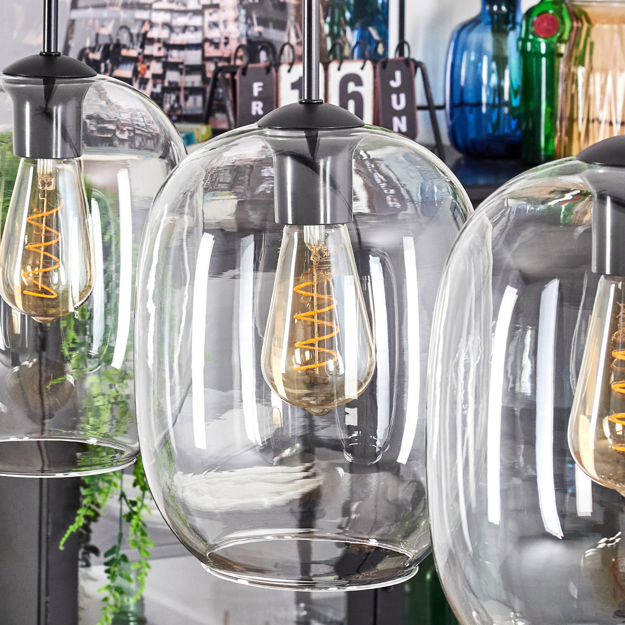 Nahaufnahme von Glas-Pendelleuchten mit Edison-Glühbirnen. Im Hintergrund Flaschen und ein Kalender.