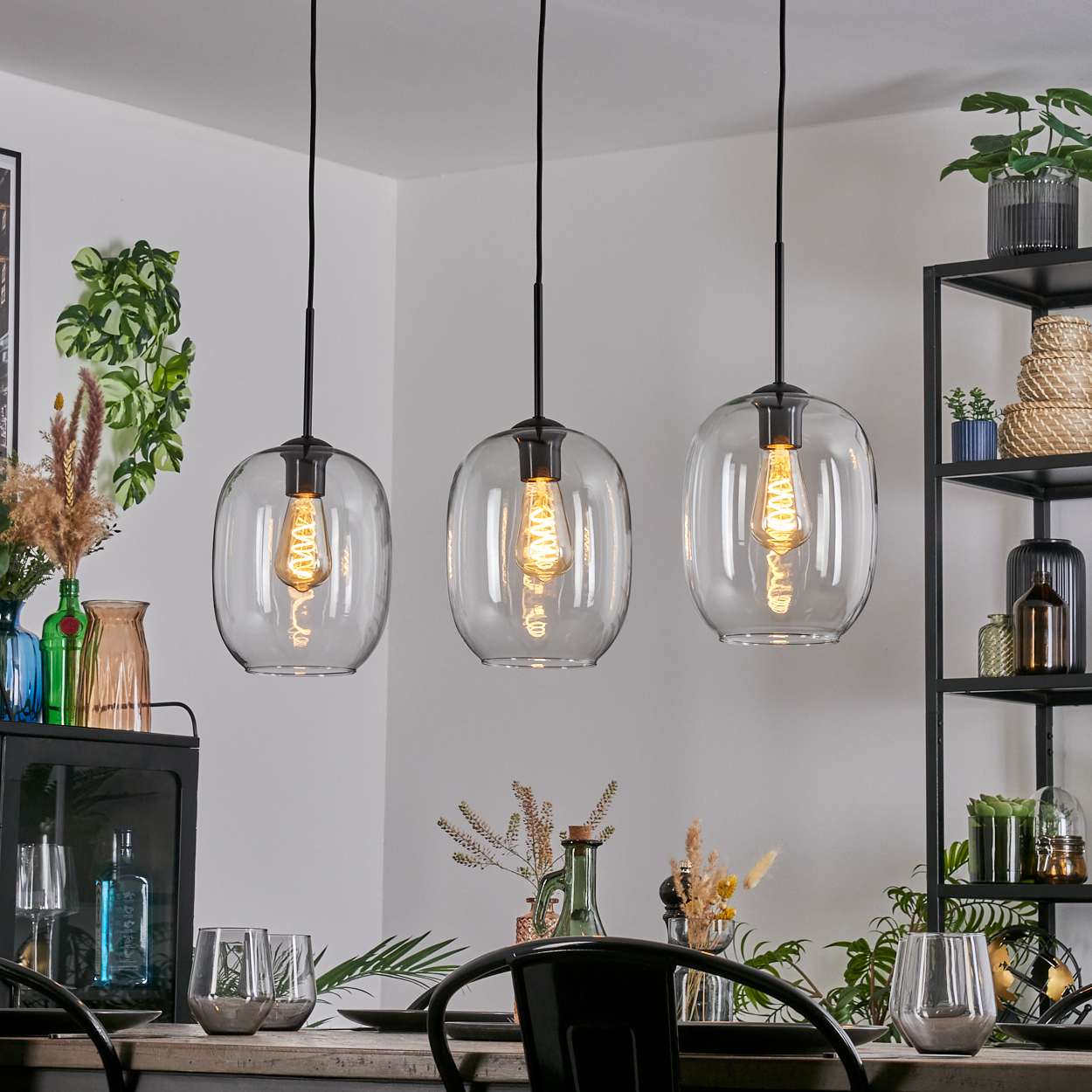 Drei Glas-Pendelleuchten hängen über einem Esstisch. Die Szene umfasst ein schwarzes Metallregal, Pflanzen und Glaswaren.
