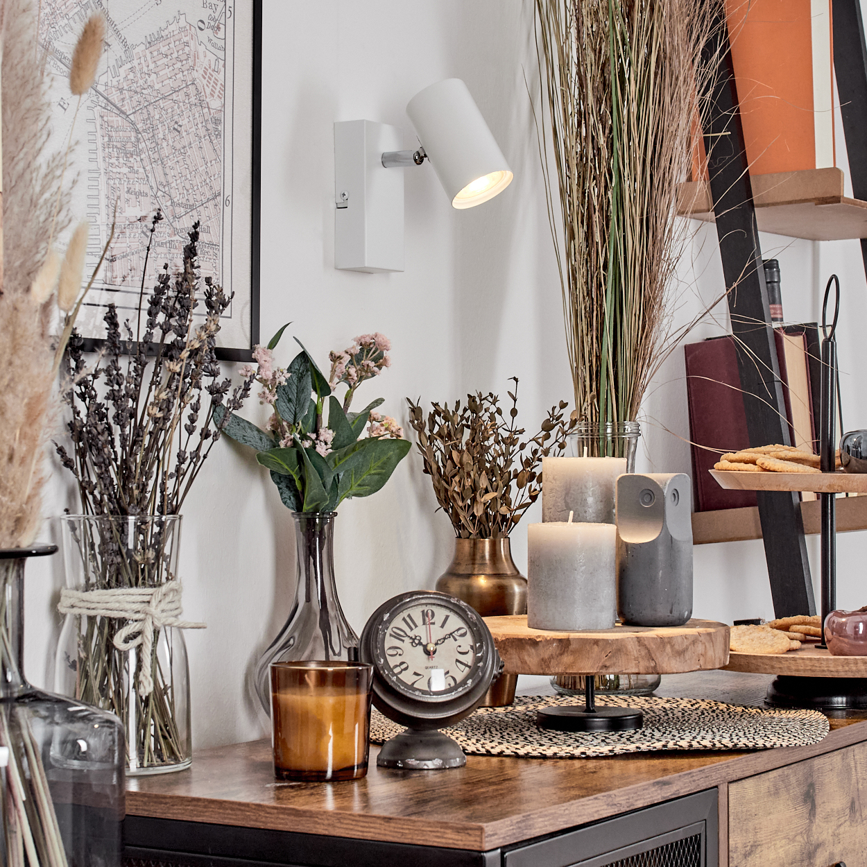 Eine dekorative Szene mit getrockneten Blumen, Kerzen, einer Uhr und einer Karte auf einer Holzoberfläche mit einer Wandlampe.