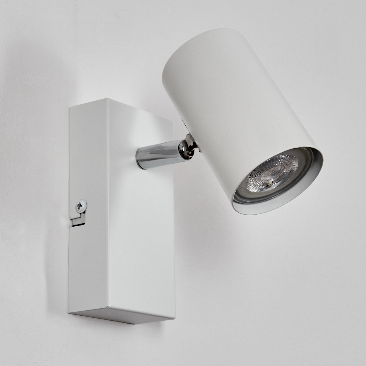Ein weißer, an der Wand montierter Strahler mit zylindrischem Kopf und rechteckiger Basis. Er hat einen Chromarm und ist vor einer weißen Wand.