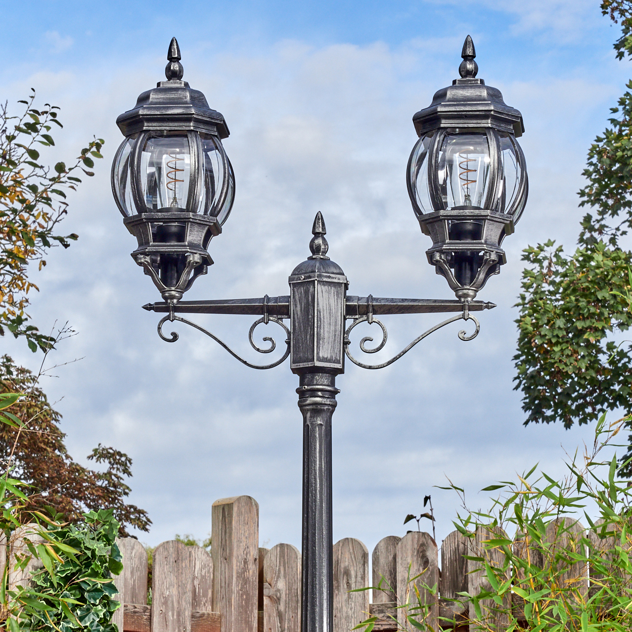 Eine dekorative Laterne mit zwei Lampen. Ein Holzzaun und grüne Pflanzen befinden sich im Vordergrund, mit blauem Himmel im Hintergrund.