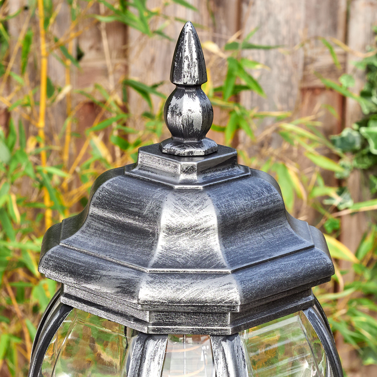Nahaufnahme einer dekorativen Außenlampe mit silberfarbener Oberseite und Glasscheiben. Bambus und ein Holzzaun befinden sich im Hintergrund.