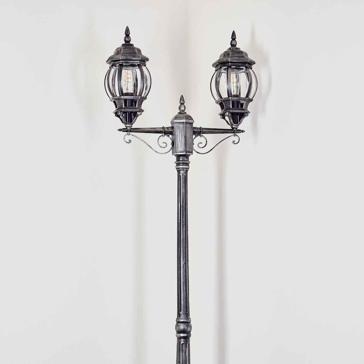 Eine verzierte Laterne mit zwei Lampen vor einer weißen Wand. Die Lampen haben klare Glaskugeln, der Pfosten ist metallisch grau.