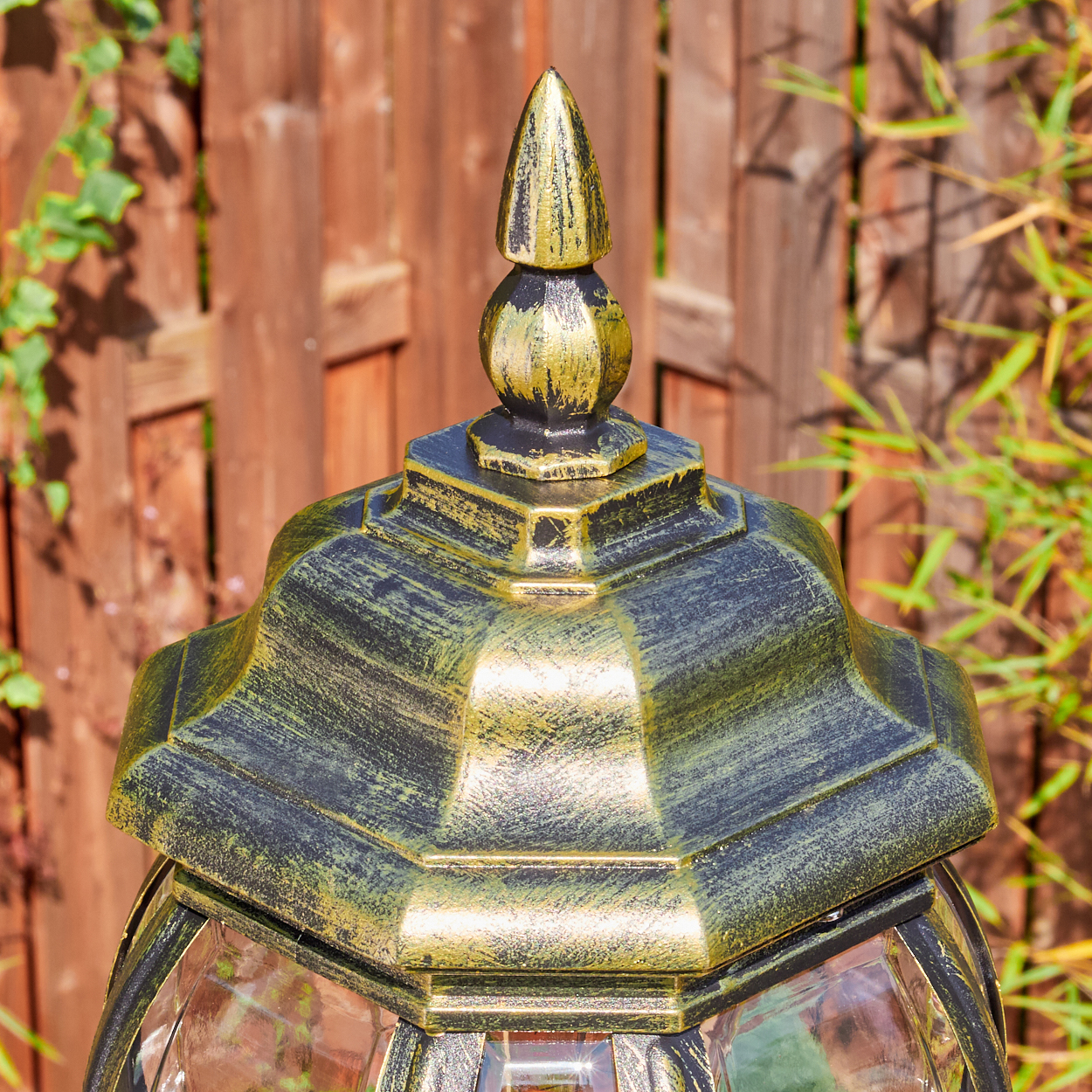 Nahaufnahme einer dekorativen Lampe mit goldener Spitze und schwarzen Akzenten. Im Hintergrund ein Holzzaun und Grün.