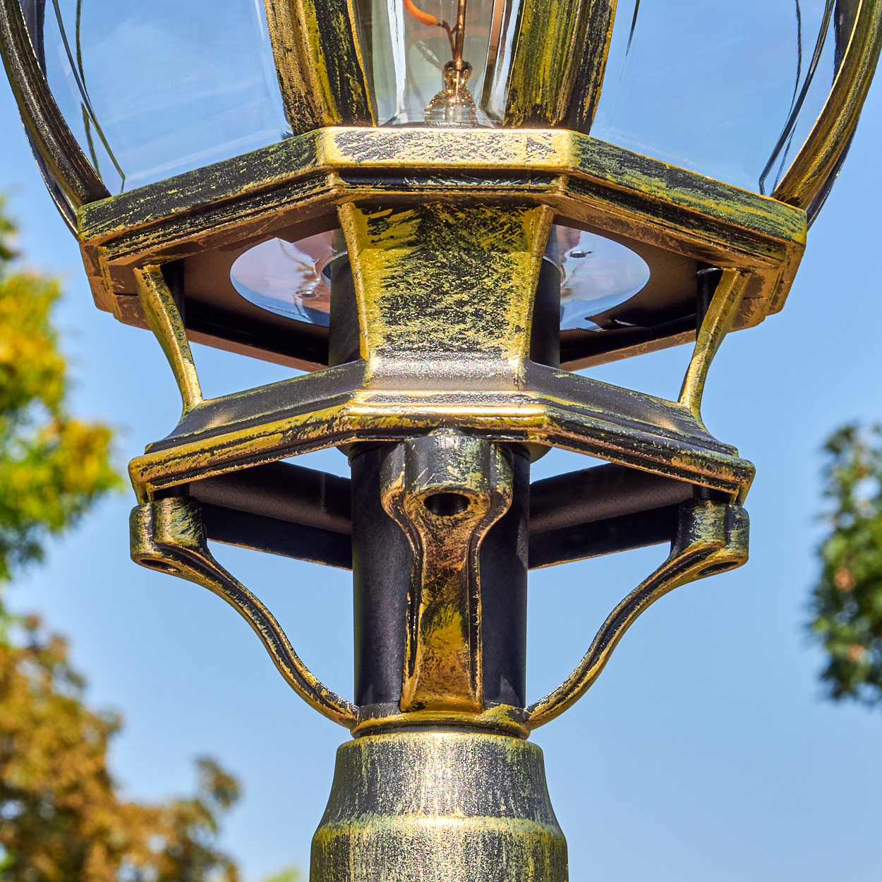 Nahaufnahme einer dekorativen Laterne mit goldenen und schwarzen Details vor blauem Himmel, sichtbaren Glasscheiben und einer Glühbirne.
