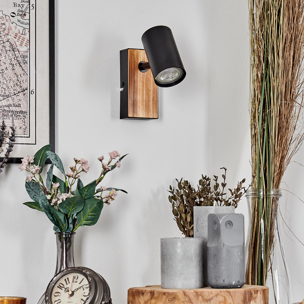 Eine moderne Wandlampe mit Holzsockel und schwarzem Lampenkopf. Dekorative Pflanzen und eine Eulenstatue stehen darunter. Im Hintergrund ist eine Karte zu sehen.