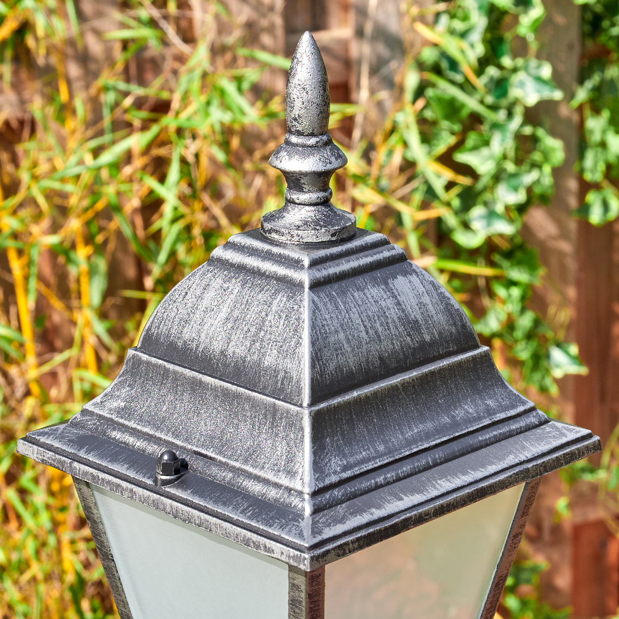 Nahaufnahme einer silbernen Außenlampe. Der Hintergrund zeigt grüne Pflanzen und einen Holzzaun, Sonnenlicht ist sichtbar.