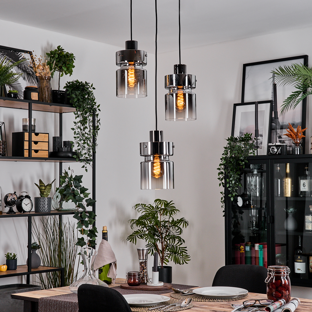 Esszimmer mit drei Hängelampen. Tisch mit Tellern und Besteck. Regale und Schrank mit Pflanzen und Dekorationsartikeln.