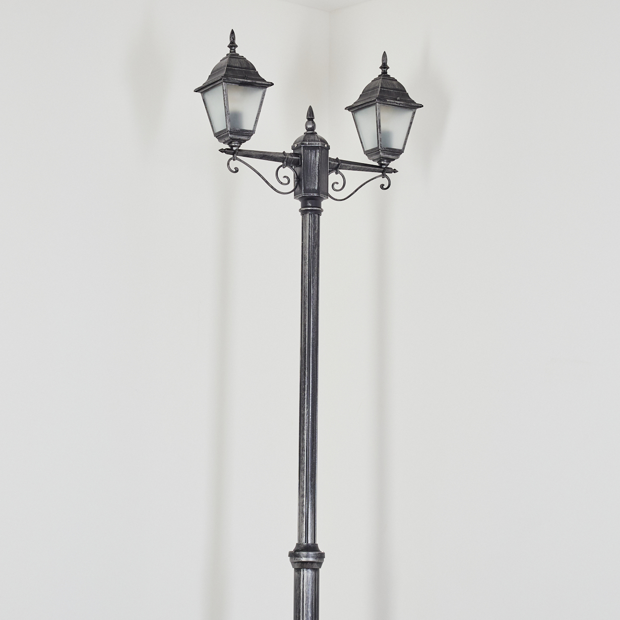 Eine dekorative, antike Straßenlaterne mit zwei Lichtern. Die Stange ist dunkelgrau, vor einer weißen Wand und Ecke.