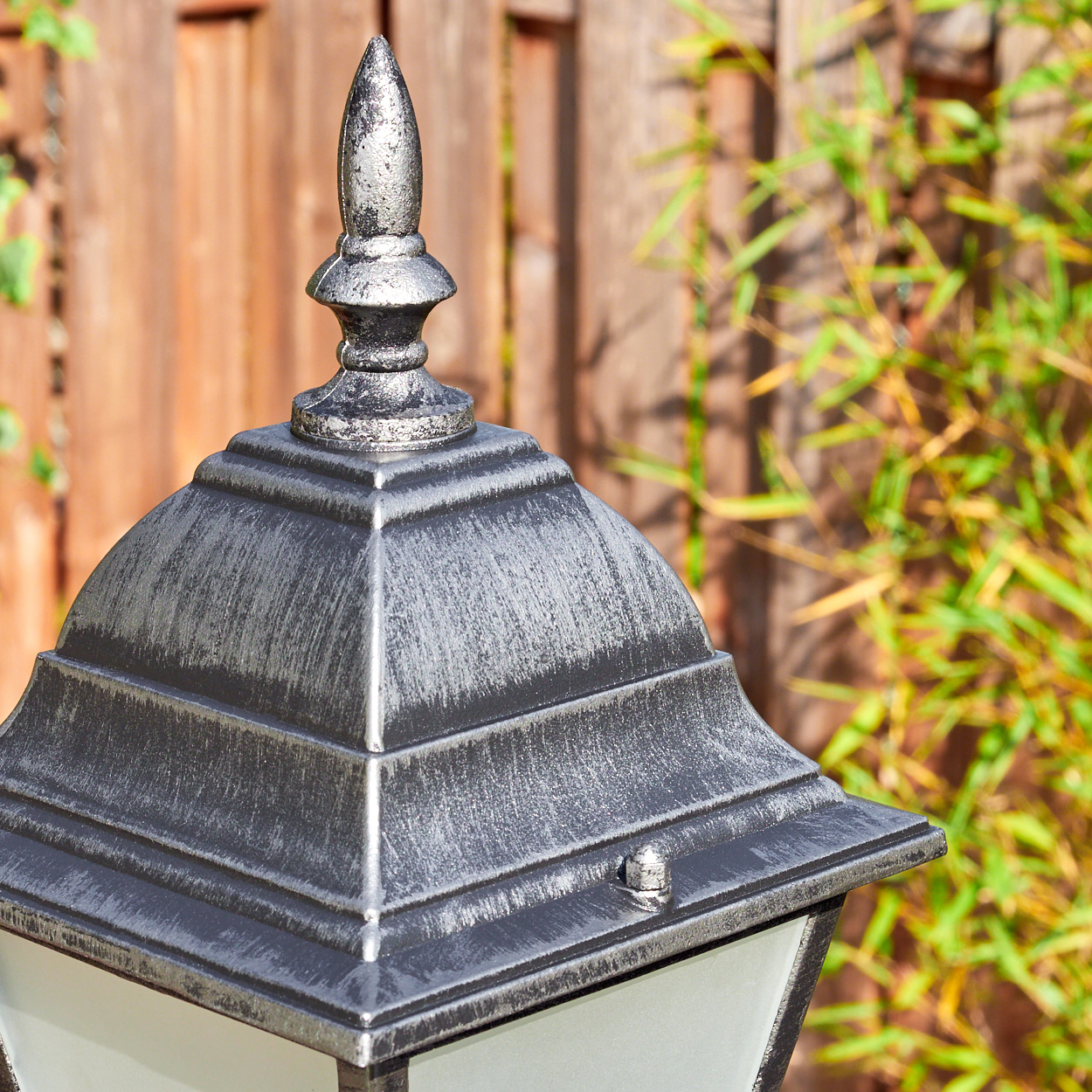 Nahaufnahme einer silbernen Außenlampe. Die Lampe hat eine dekorative Spitze, im Hintergrund ein Holzzaun und Bambus.