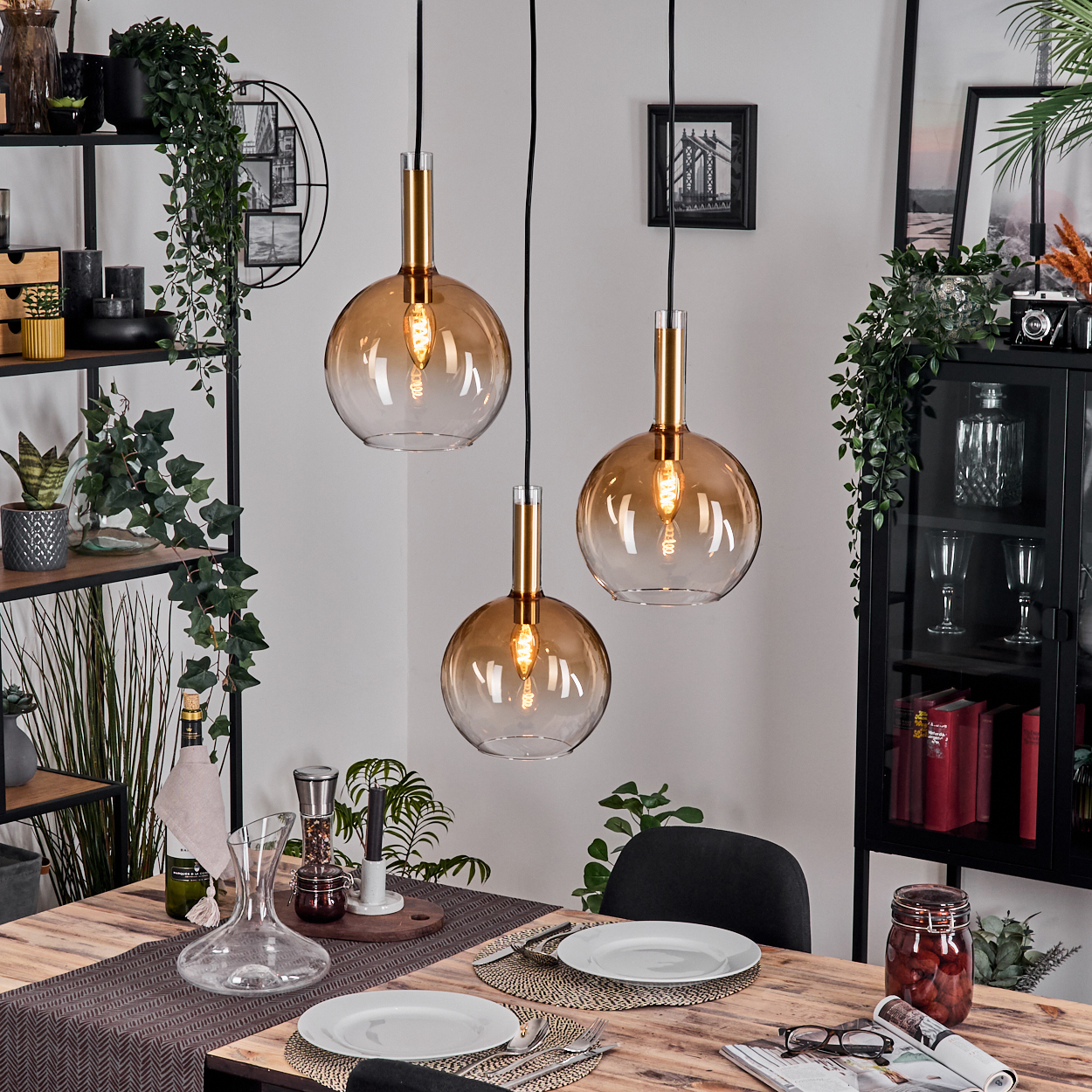 Drei Pendelleuchten über einem Holztisch, der für das Abendessen gedeckt ist. Pflanzen und Dekorationsartikel umgeben den Tisch.