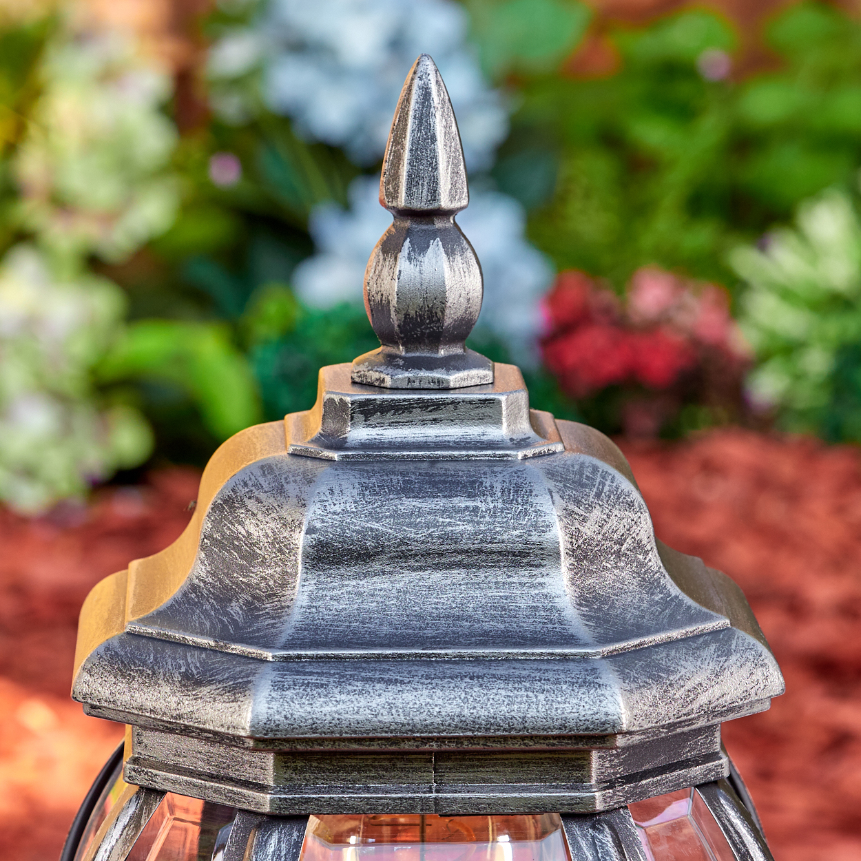 Nahaufnahme einer dekorativen Lampe mit Metallic-Finish, vor verschwommenem Hintergrund aus roter Mulch und grünen Pflanzen.