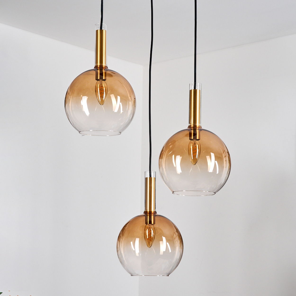 Drei Pendelleuchten mit bernsteinfarbenen Glasschirmen und goldenen Akzenten hängen an einer weißen Wand. Schwarze Kabel halten sie.