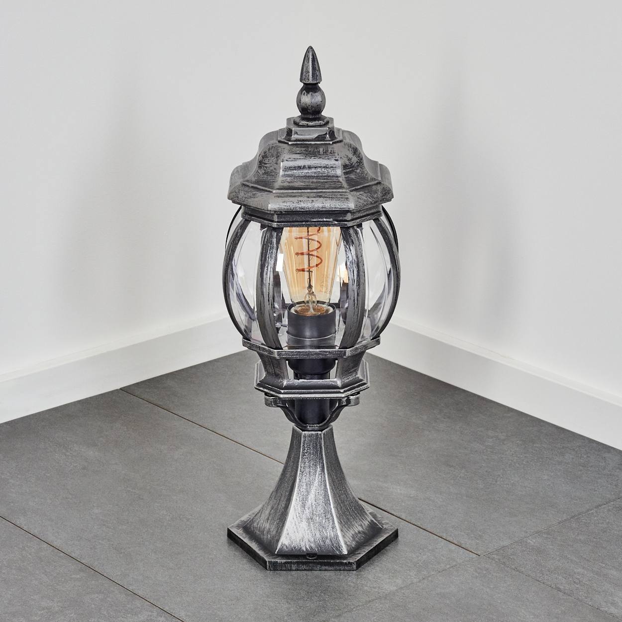 Eine dekorative Außenlampe, silber und schwarz, mit einem klaren Glasgehäuse für die Glühbirne, auf einem grauen Fliesenboden platziert.