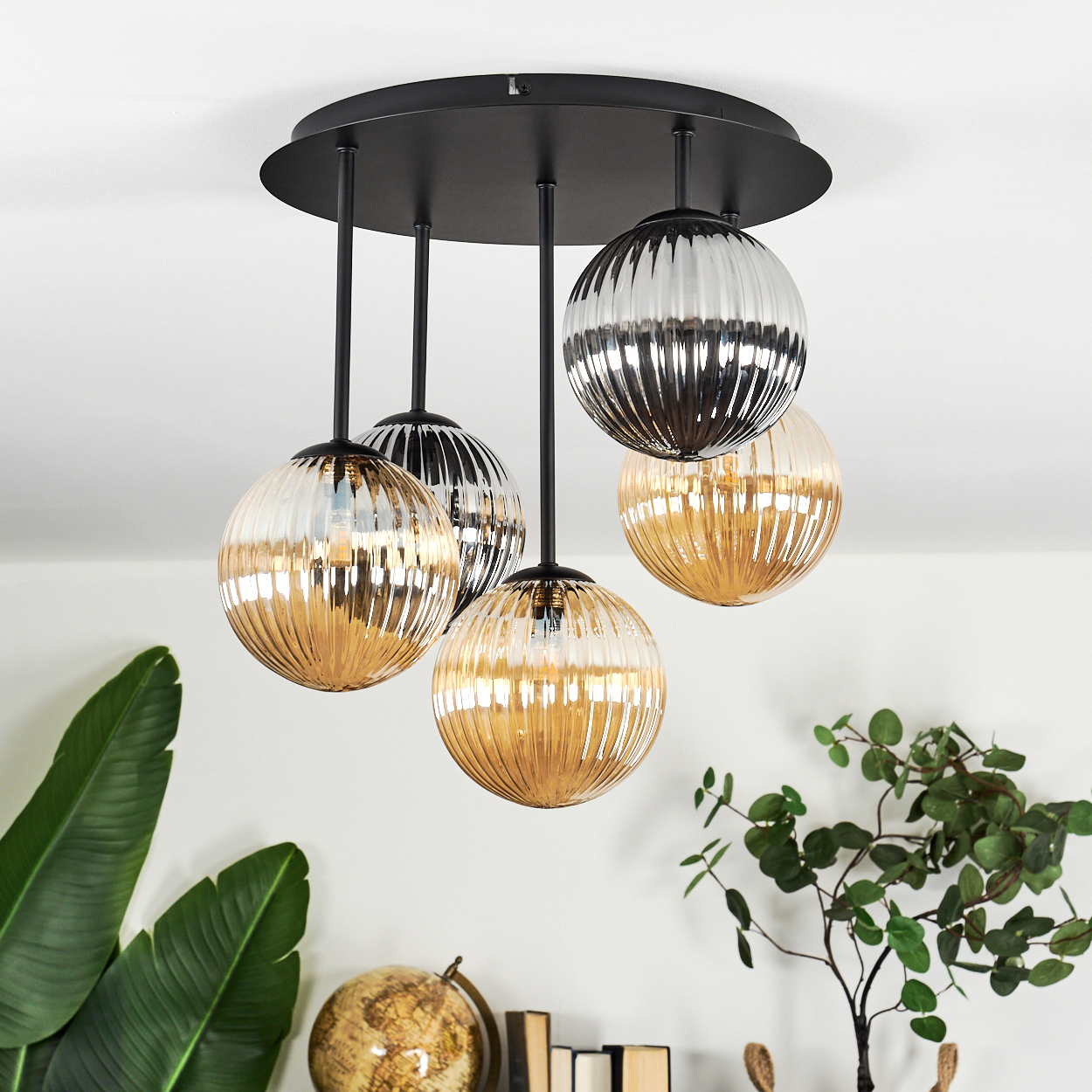 Moderne Deckenleuchte mit fünf kugelförmigen Lampen. Die Lampen haben eine Gold- und Silberoberfläche mit schwarzem Sockel. Pflanzen und Globus im Hintergrund.