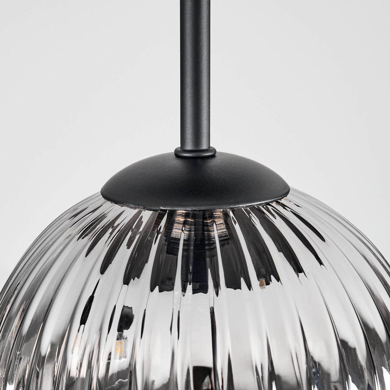 Nahaufnahme einer Glas-Pendelleuchte mit geripptem Design. Die Leuchte hat einen schwarzen Metallaufsatz und einen durchsichtigen, gewölbten Schirm. Der Hintergrund ist weiß.