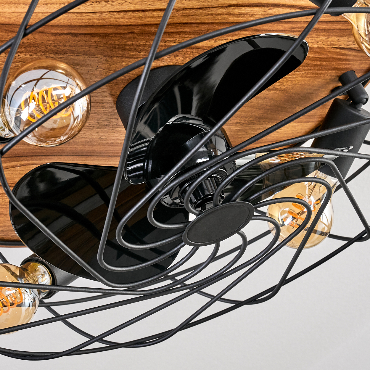 Nahaufnahme eines Deckenventilators mit schwarzen Flügeln und einem Drahtkäfig. Mehrere Glühbirnen sind auf einem Holzgrund sichtbar.
