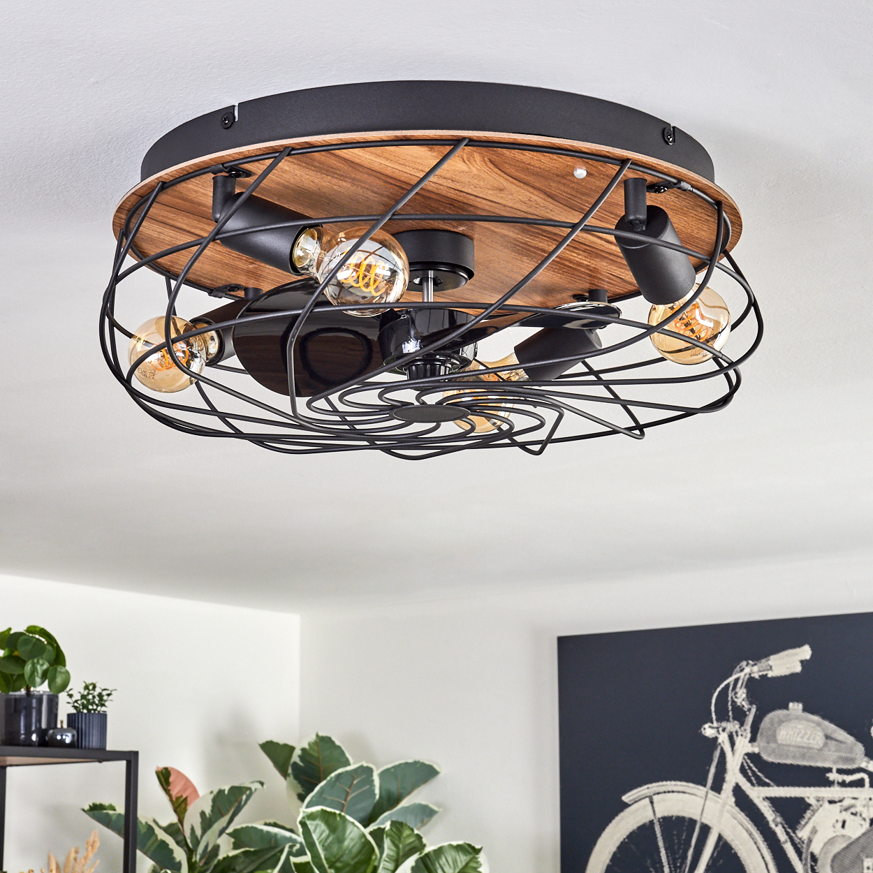 Eine Deckenleuchte mit freiliegenden Glühbirnen und einem schwarzen Metallkäfig. Holzähnliche Basis und schwarzer Ventilator. Pflanzen und ein Fahrradkunstwerk im Hintergrund.