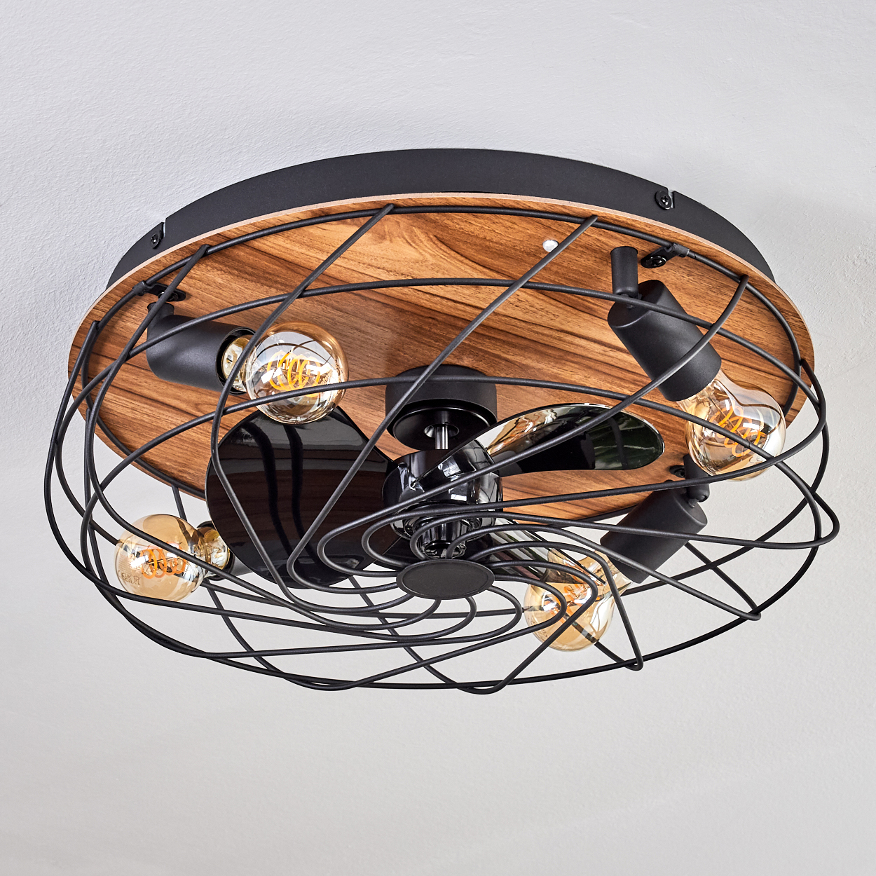 Ein Deckenventilator mit freiliegenden Glühbirnen. Er hat einen Holzsockel, einen schwarzen Metallkäfig und einen schwarzen Ventilator. Die Glühbirnen leuchten.