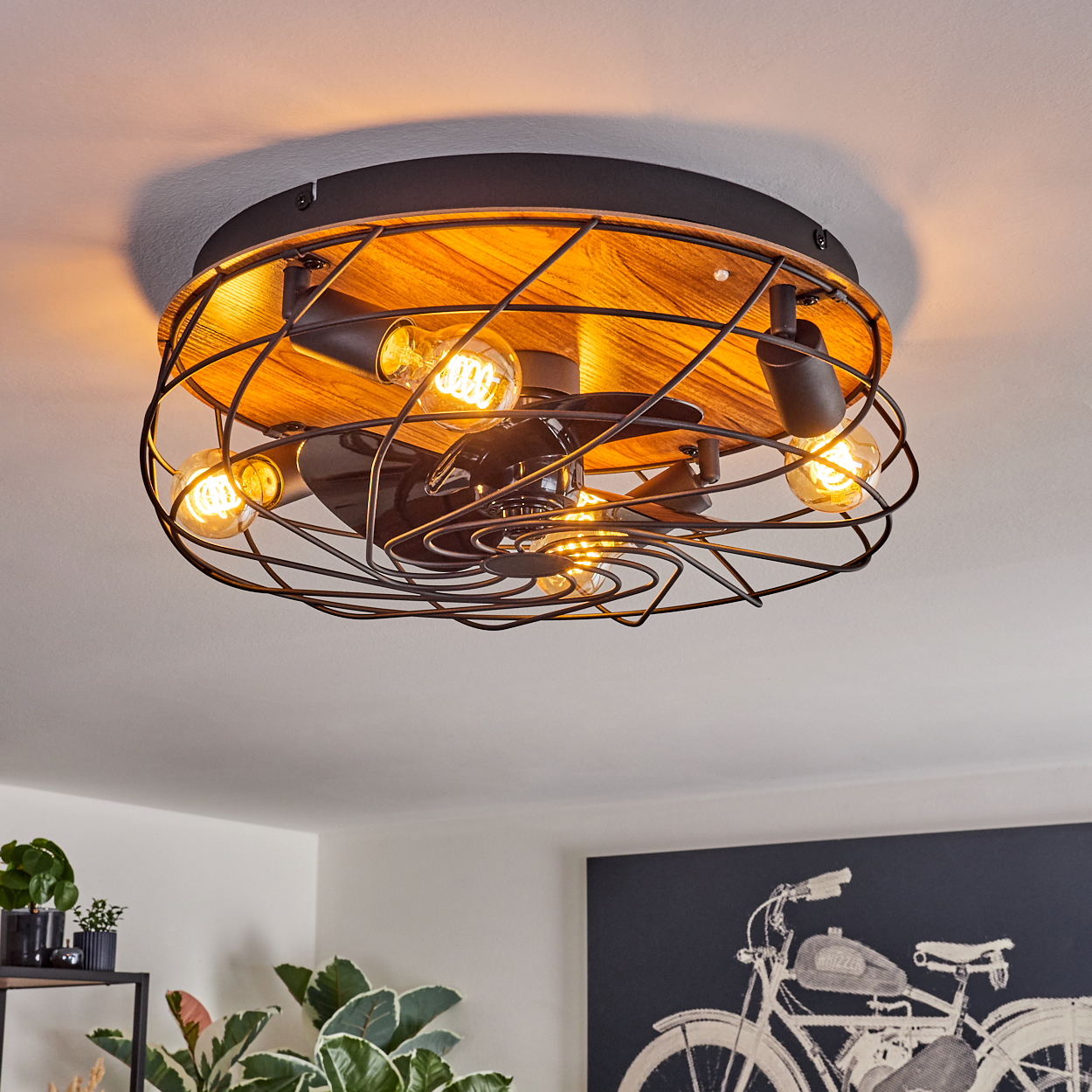 Deckenventilator mit freiliegenden Glühbirnen und Holzsockel. Der Ventilator ist schwarz, die Glühbirnen leuchten. Der Hintergrund zeigt eine Raumumgebung.