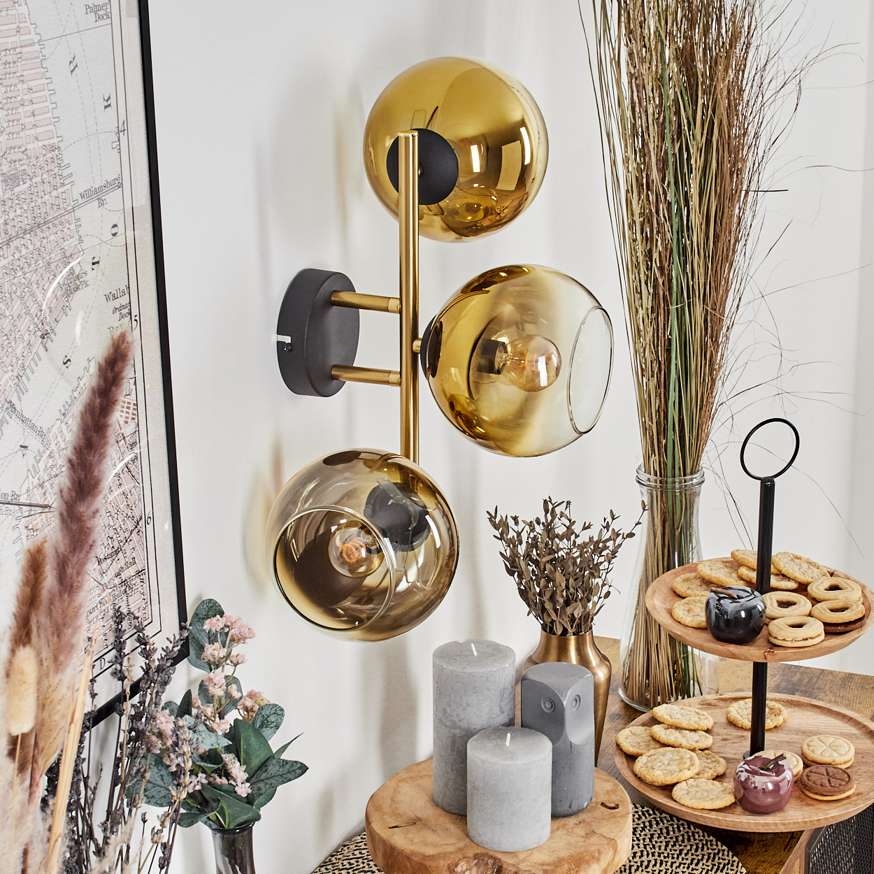 Eine moderne Wandleuchte mit drei goldenen Glaskugeln, Kerzen, Keksen auf einem Holzständer und Zierpflanzen.