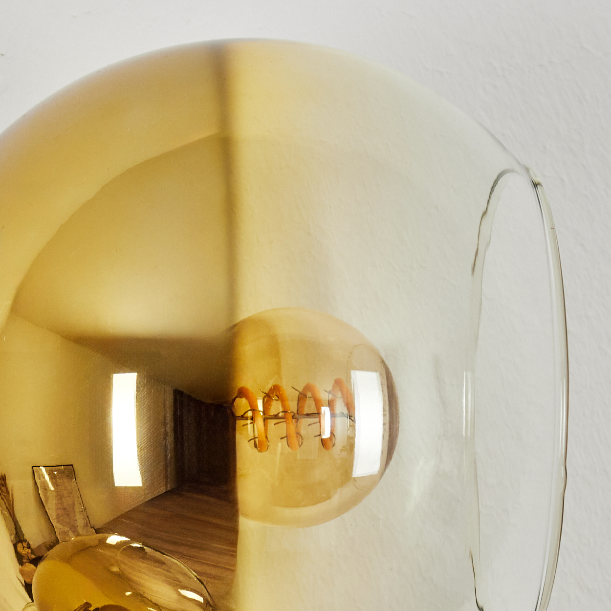 Nahaufnahme einer Glühbirne in einem Glasschirm. Die Glühbirne hat einen spiralförmigen Glühfaden. Der Hintergrund ist ein Raum mit Holzboden.