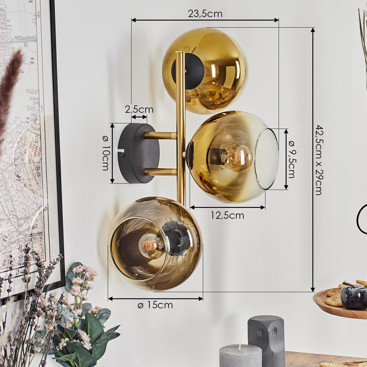 Eine moderne Wandlampe mit drei goldenen Glaskugelschirmen. Maße und eine Karte sind im Hintergrund sichtbar. Weitere Dekorationen sind ebenfalls vorhanden.