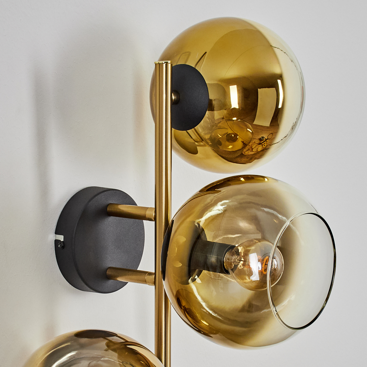 Eine moderne Wandlampe mit drei goldenen Glaskugel-Schirmen und einem schwarzen Sockel, an einer weißen Wand befestigt.