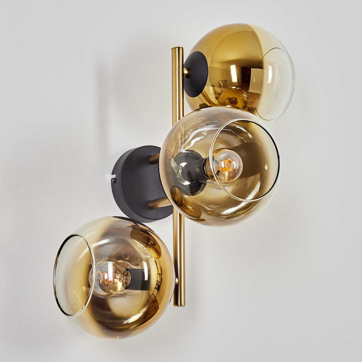 Eine Wandlampe mit drei goldfarbenen Glaskugeln, die jeweils eine Glühbirne enthalten, an einer weißen Wand befestigt.