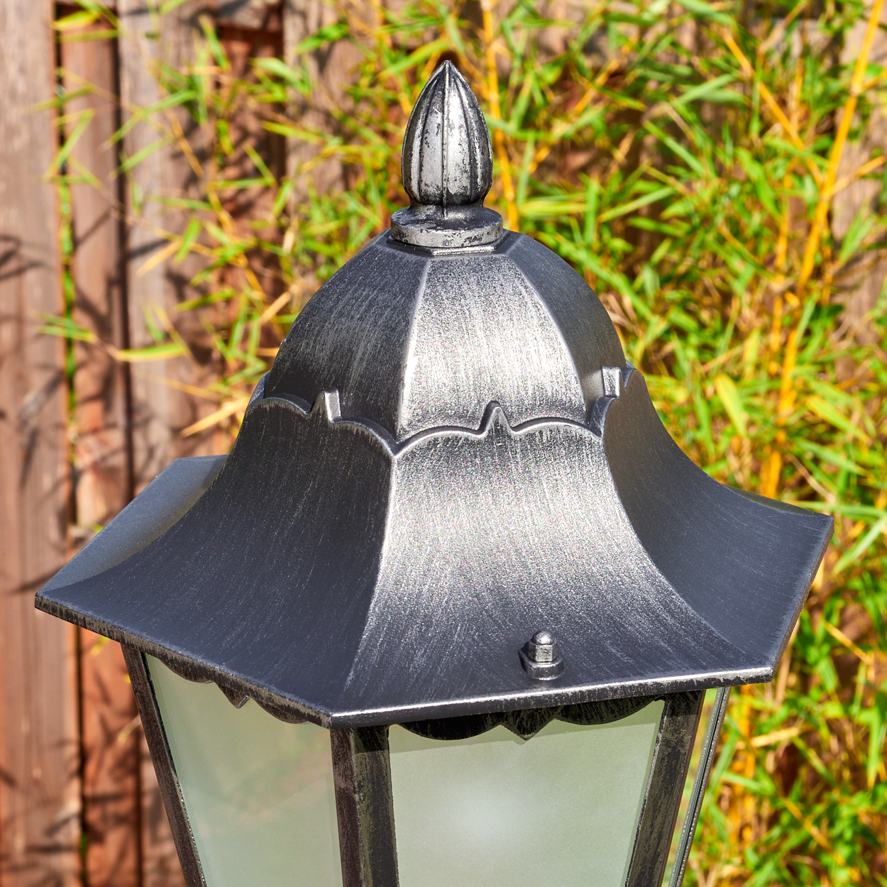 Nahaufnahme einer dekorativen Lampe mit metallgrauem Finish, vor einem Holzzaun und grünem Bambus.