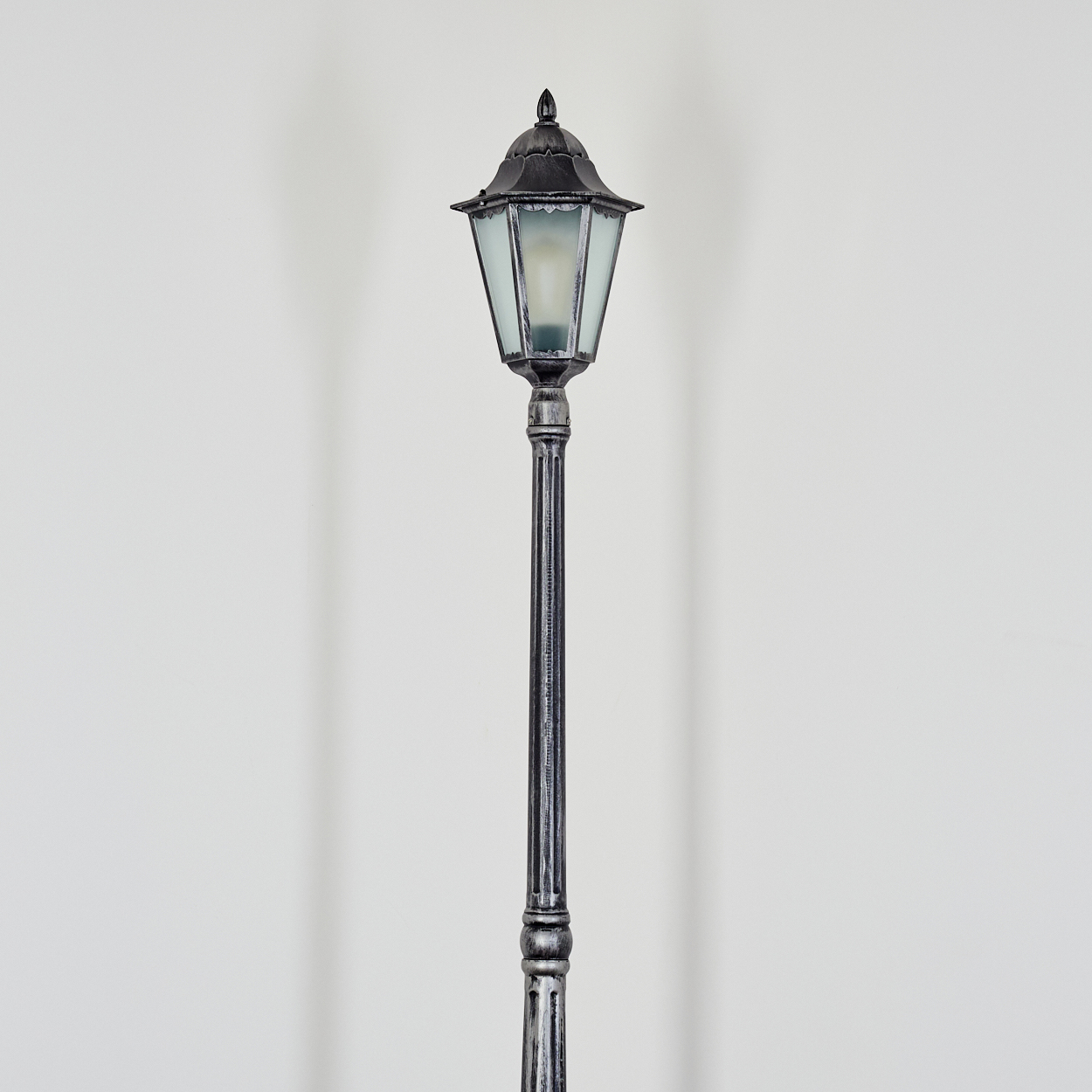Eine dekorative, schwarz-silberne Straßenlaterne steht vor einer weißen Wand. Die Lampe hat einen Glasschirm und verzierte Details.