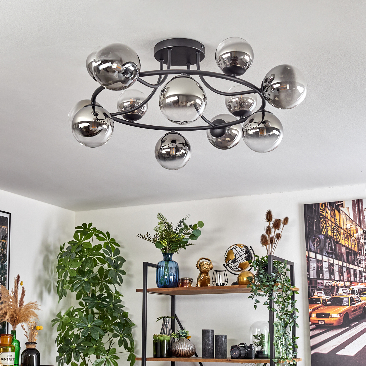 Eine moderne Deckenleuchte mit reflektierenden Kugeln. Darunter ein Regal mit Pflanzen, Dekoration und einem gerahmten Stadtbild.