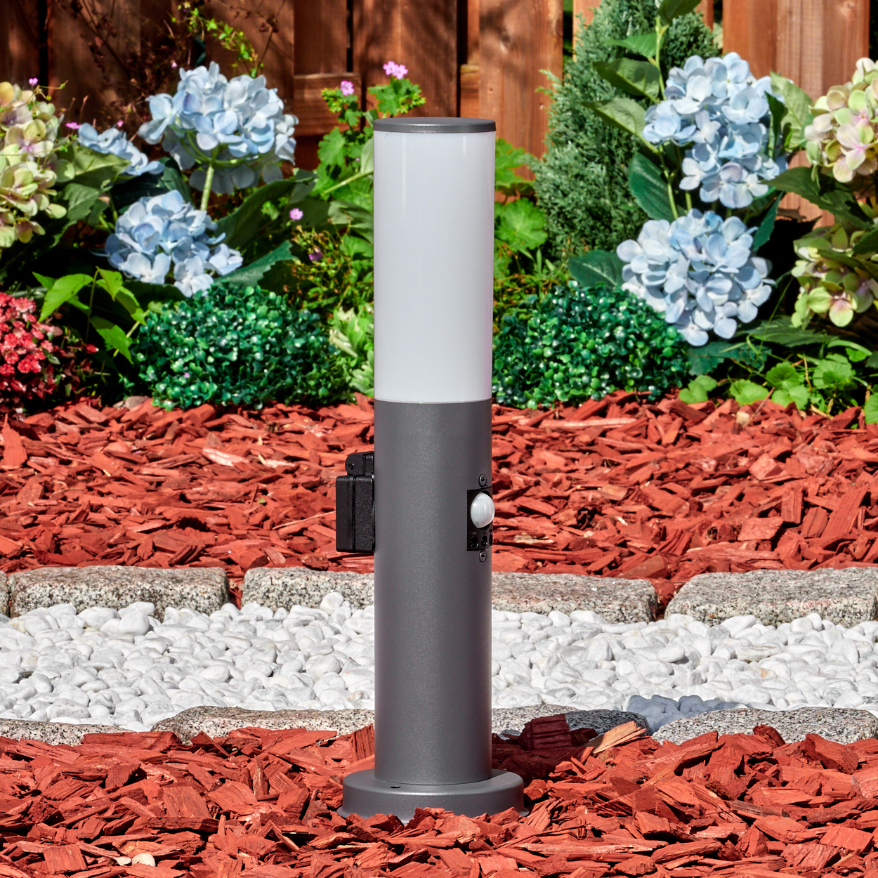 Eine graue Außenlampe steht in einem Gartenbeet mit roter Mulch, weißen Steinen und blauen Blumen. Im Hintergrund ist ein Holzzaun.