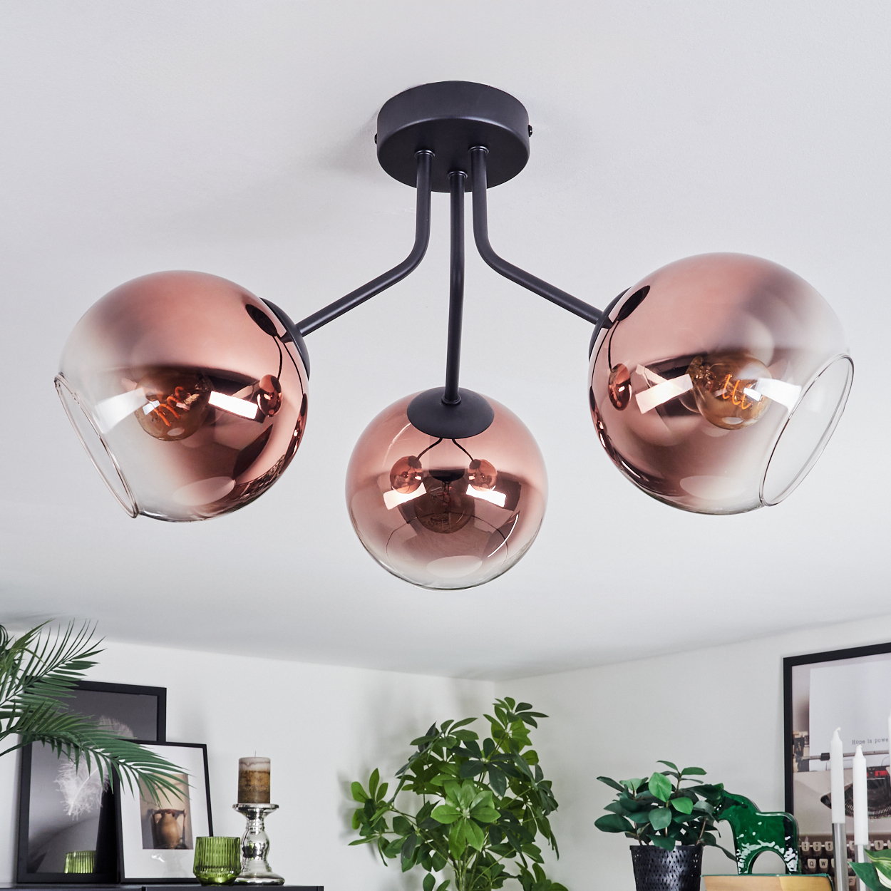 Eine moderne Deckenlampe mit drei kugelförmigen Schirmen in Roségold und klarem Glas, die in einem Raum mit Pflanzen hängt.