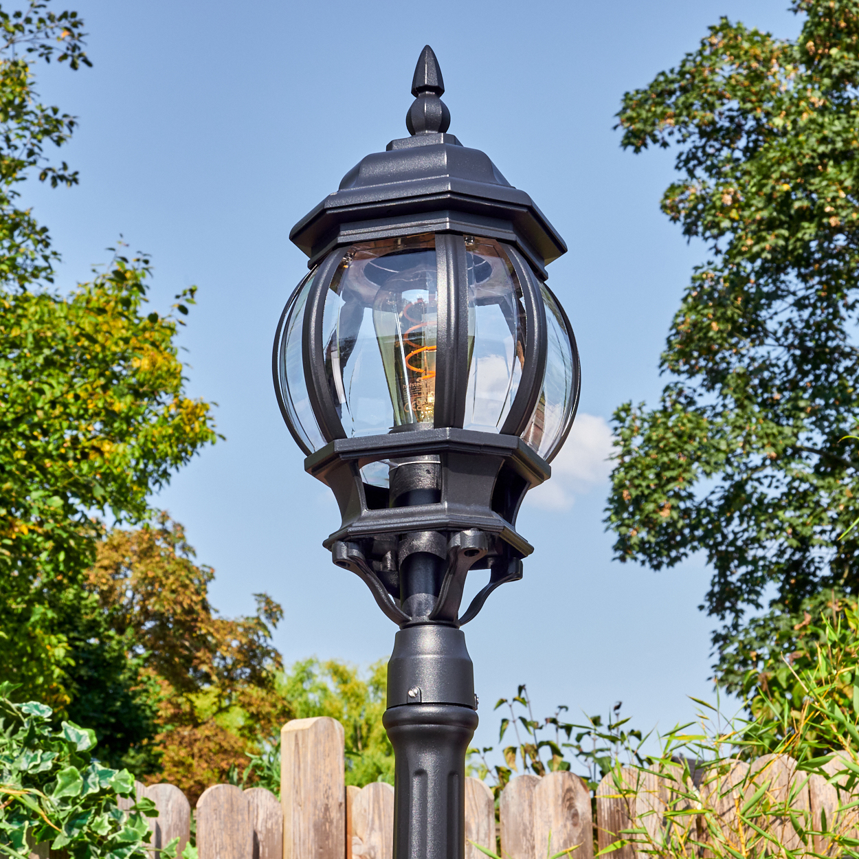 Schwarze Außenlampe mit Glühbirne. Sie befindet sich in einem Garten mit Holzzaun und grünen Bäumen unter blauem Himmel.