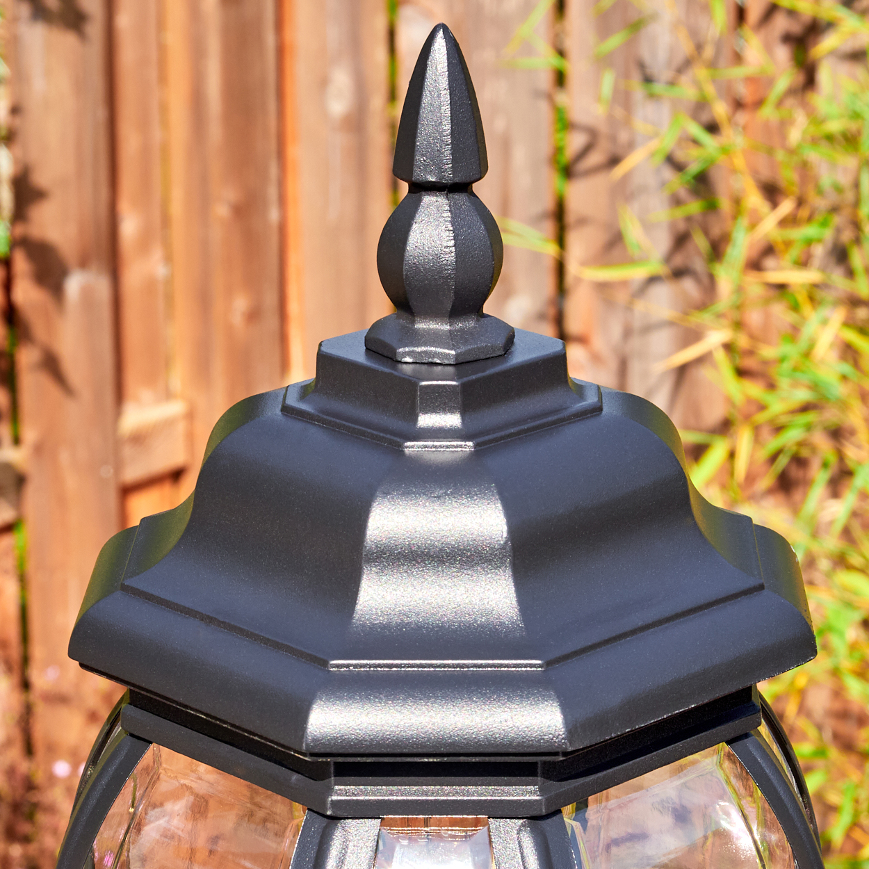 Nahaufnahme einer schwarzen Außenlampe mit spitzer Spitze. Holzzaun und grüne Pflanzen im unscharfen Hintergrund.