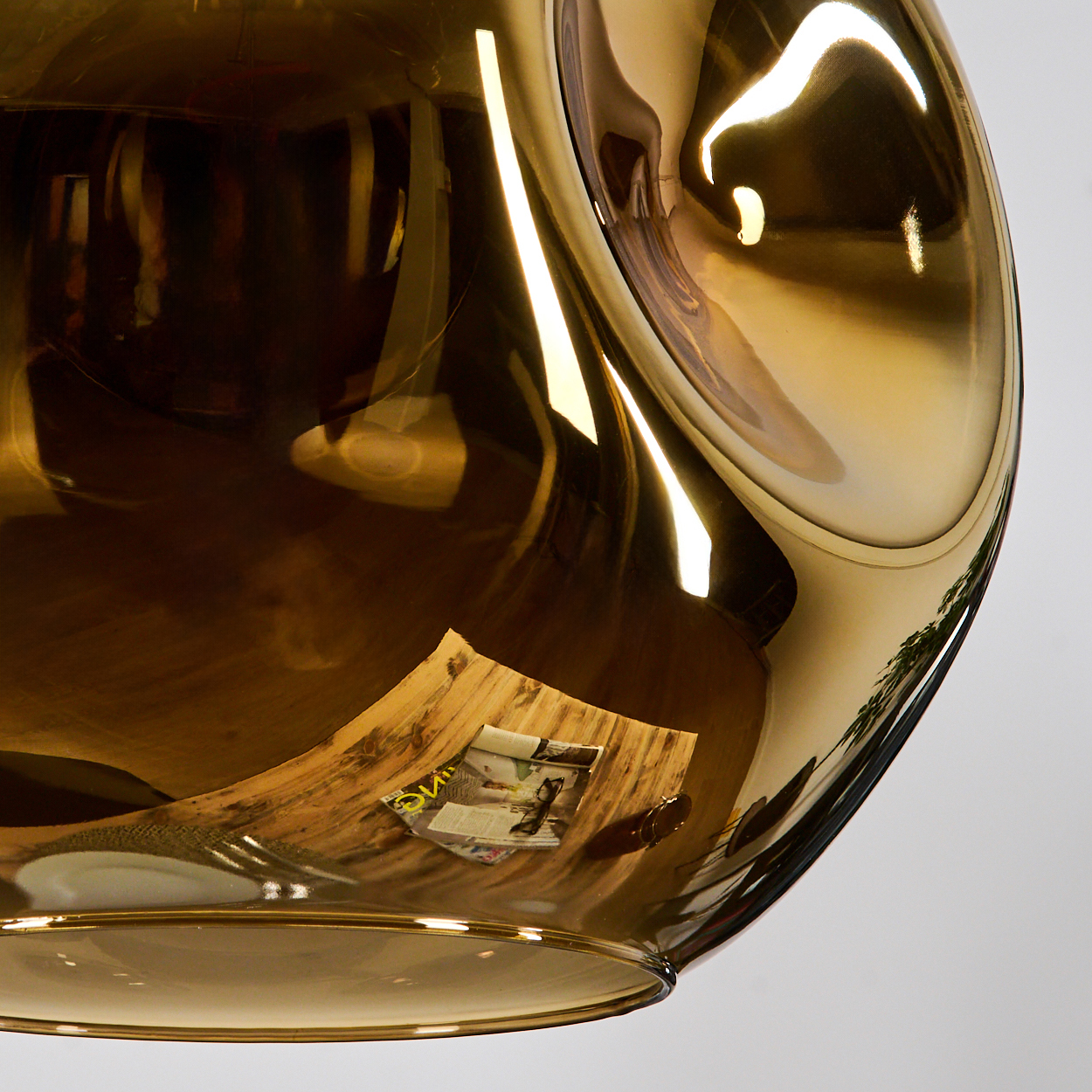 Ein goldfarbenes, tropfenförmiges Glasobjekt reflektiert eine Zeitschrift und ein Licht. Der Hintergrund ist weiß.